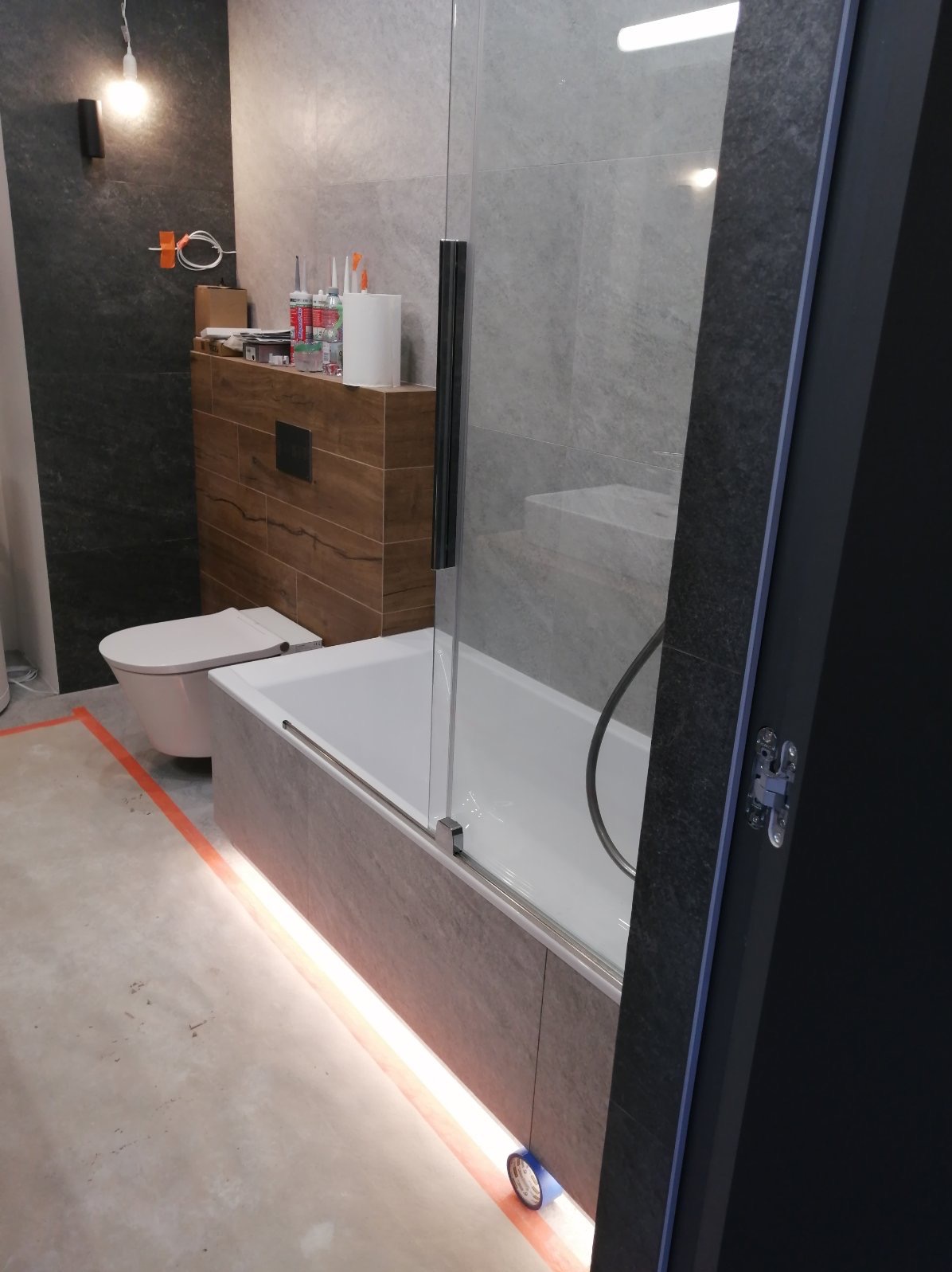 Nowoczesna łazienka w trakcie wykańczania, widoczna wanna z przeszklonym parawanem, podświetlenie LED u dołu wanny, toaleta w zabudowie, szare płytki na ścianach i podłodze częściowo zabezpieczonej...