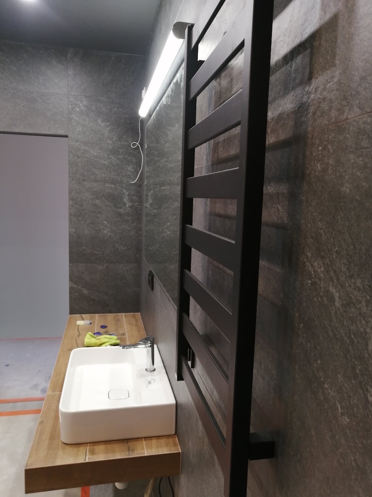 Nowoczesna łazienka z umywalką nablatową na drewnianym blacie, ciemnoszara ściana z grzejnikiem drabinkowym i podłużnym oświetleniem LED.