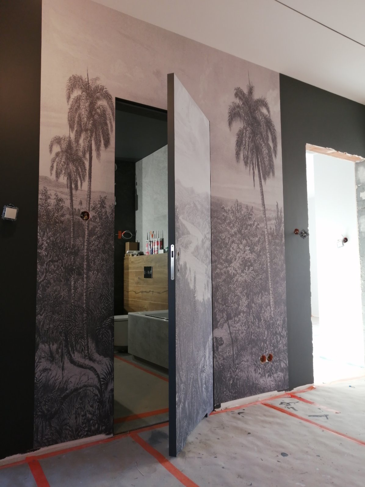 Ściana z fototapetą przedstawiającą tropikalny krajobraz z palmami, częściowo zasłonięta przez otwarte drzwi przesuwne, widoczne przygotowania do dalszych prac wykończeniowych, w tle fragment...