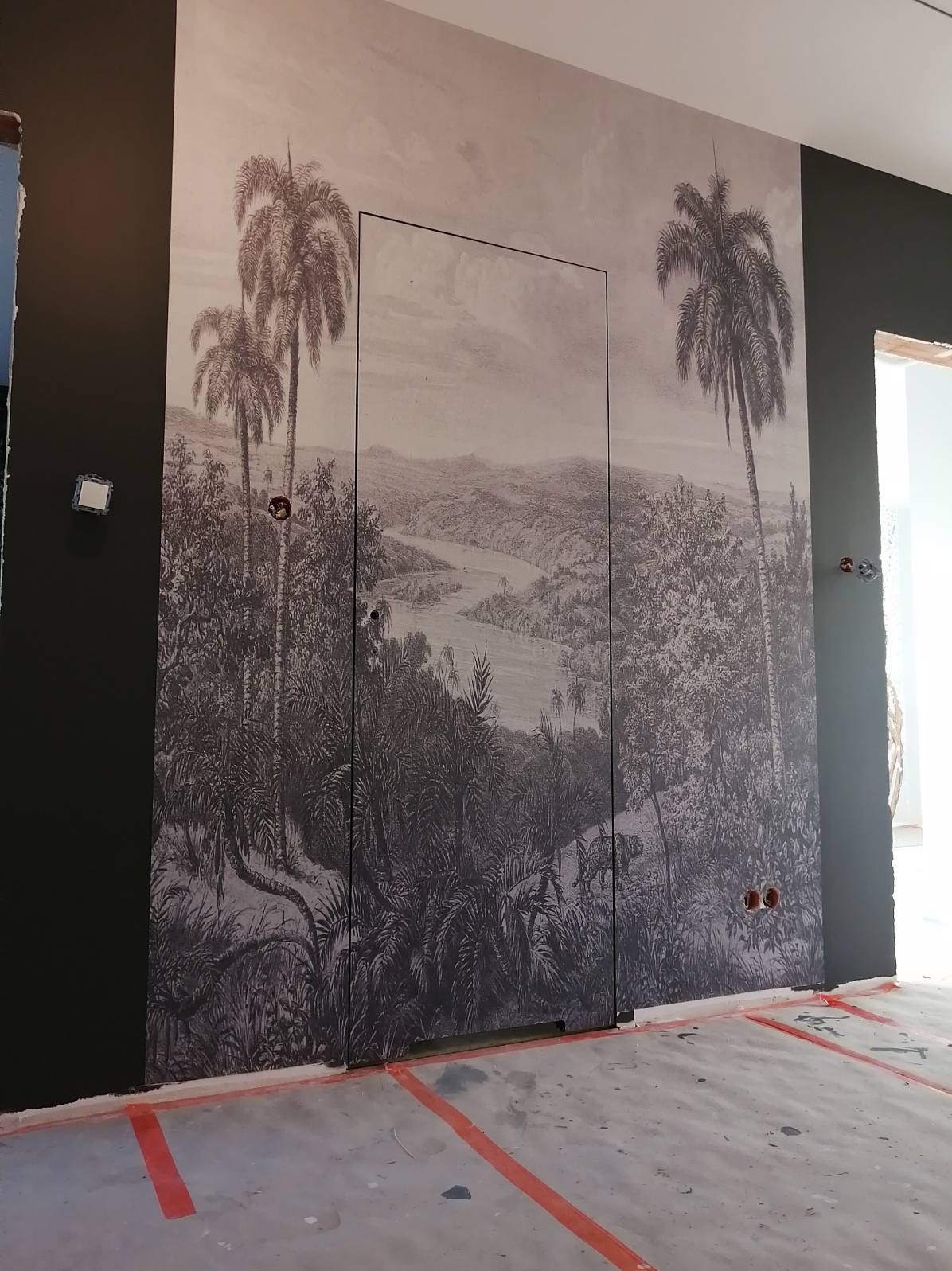 Ściana z fototapetą przedstawiającą tropikalny krajobraz, ukryte drzwi z czarną ramą, surowa podłoga z pomarańczowymi oznaczeniami.