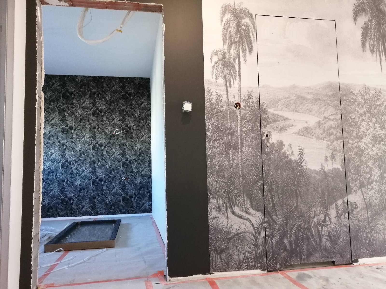 Wnętrze w trakcie prac wykończeniowych: widok przez otwór drzwiowy na pokój z tapetą w roślinny wzór, obok ściana pomalowana na ciemny kolor i ukryte drzwi z fototapetą przedstawiającą tropikalny...