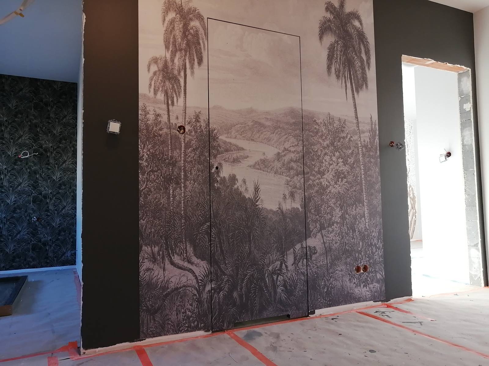Pokój w trakcie remontu z ciemną ścianą, na której umieszczono fototapetę z motywem tropikalnego krajobrazu i ukrytymi drzwiami. Podłoga zabezpieczona folią i taśmą malarską.