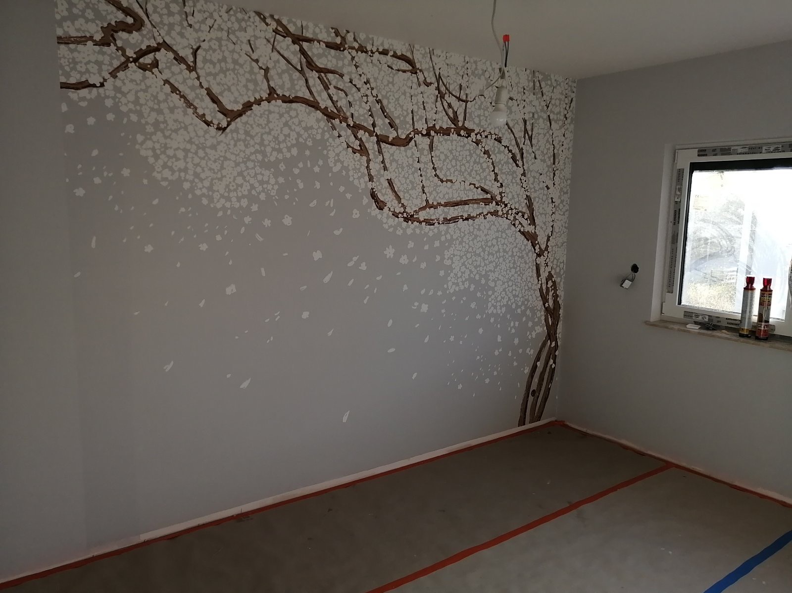 Ściana w pokoju w Warszawie z ręcznie malowanym motywem drzewa kwitnącej wiśni, widoczne puszki z farbą na parapecie okna.