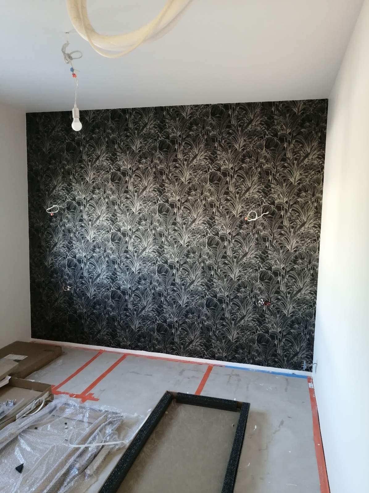 Czarno-biała tapeta w motywy roślinne na ścianie w trakcie remontu, widoczne zabezpieczenia podłogi i niedokończone prace elektryczne.