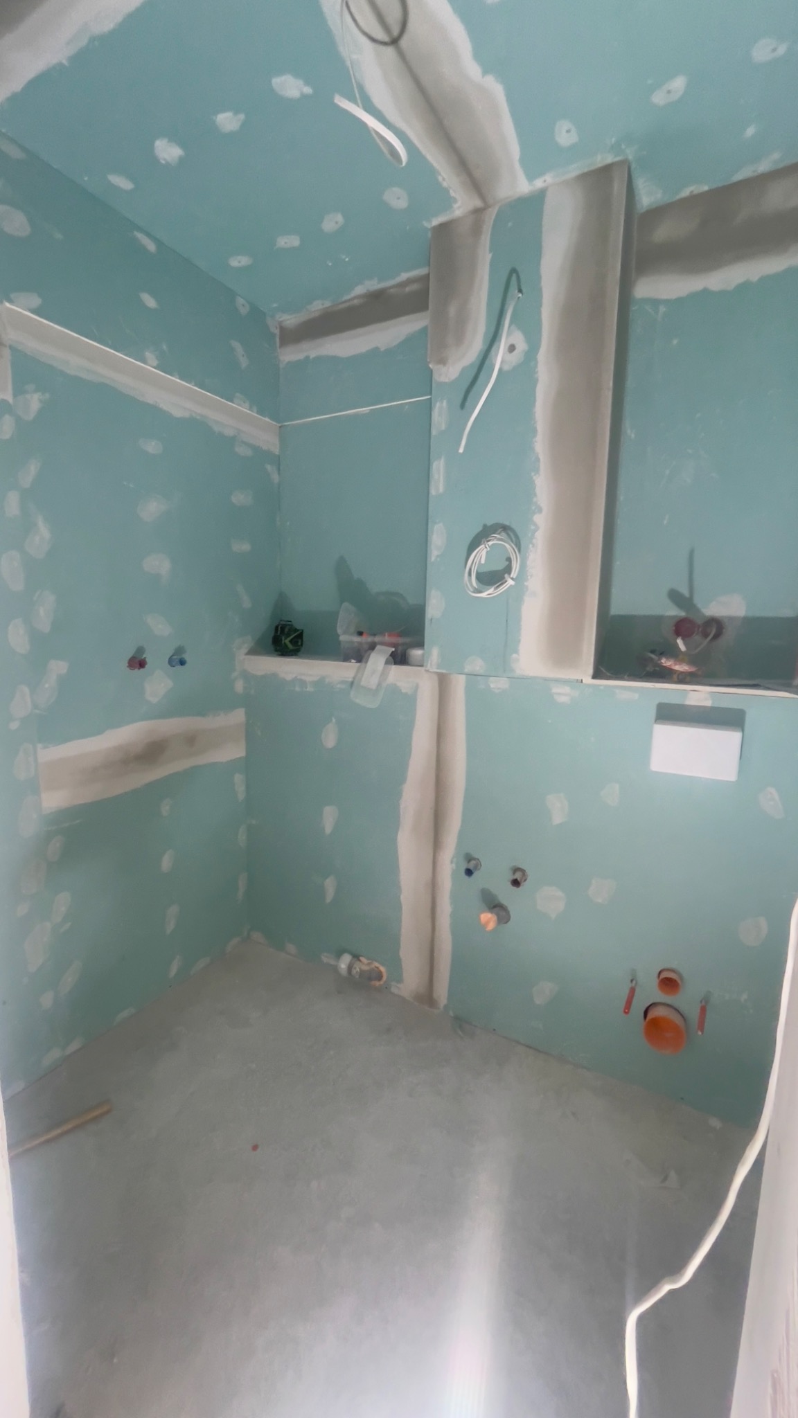 Niedokończona łazienka z płytami gipsowo-kartonowymi, widoczne instalacje wodne i elektryczne, przygotowana do dalszych prac wykończeniowych, z pozostawionymi narzędziami w tle.