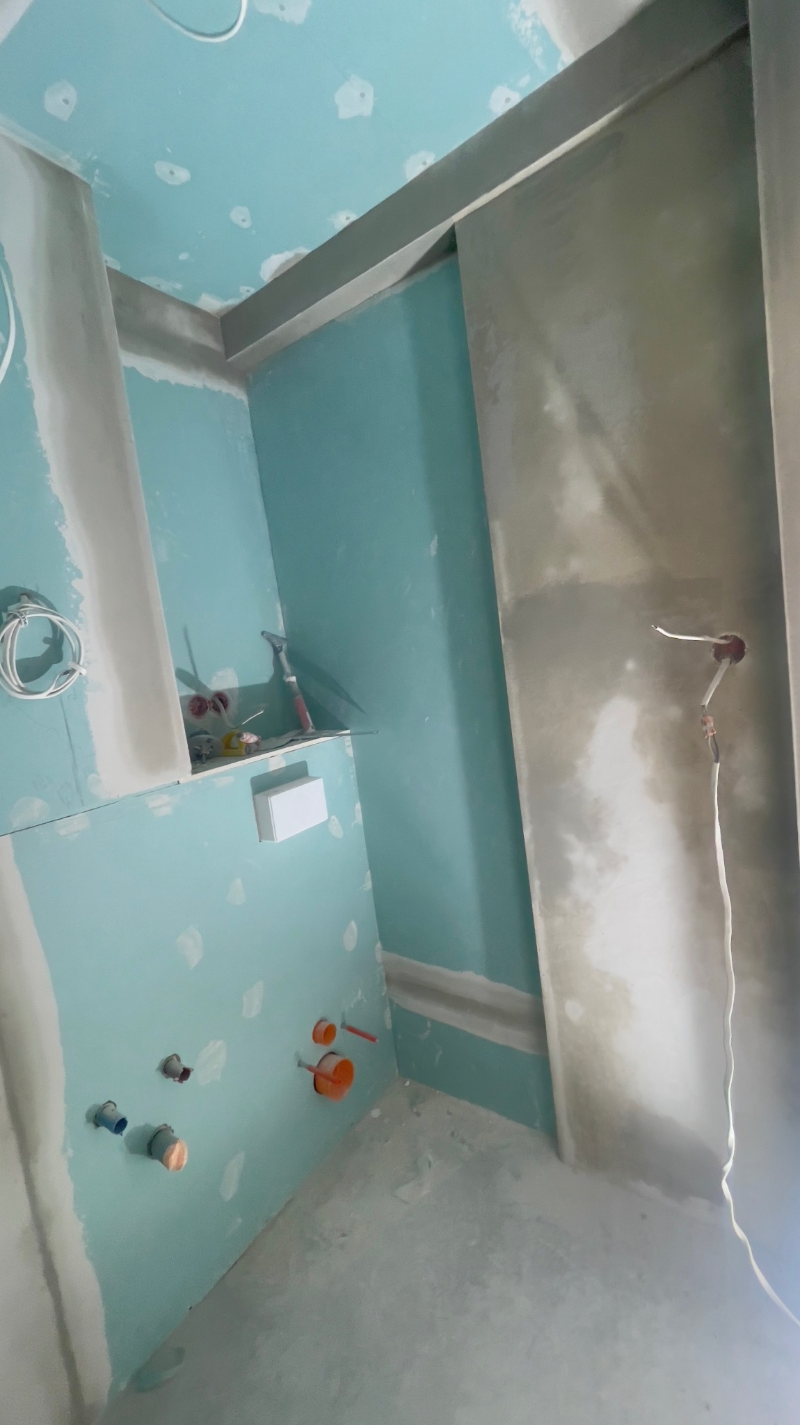 Niedokończona łazienka z płytami kartonowo-gipsowymi, widoczne instalacje wodne i elektryczne, narzędzia na półce, surowe wykończenie.
