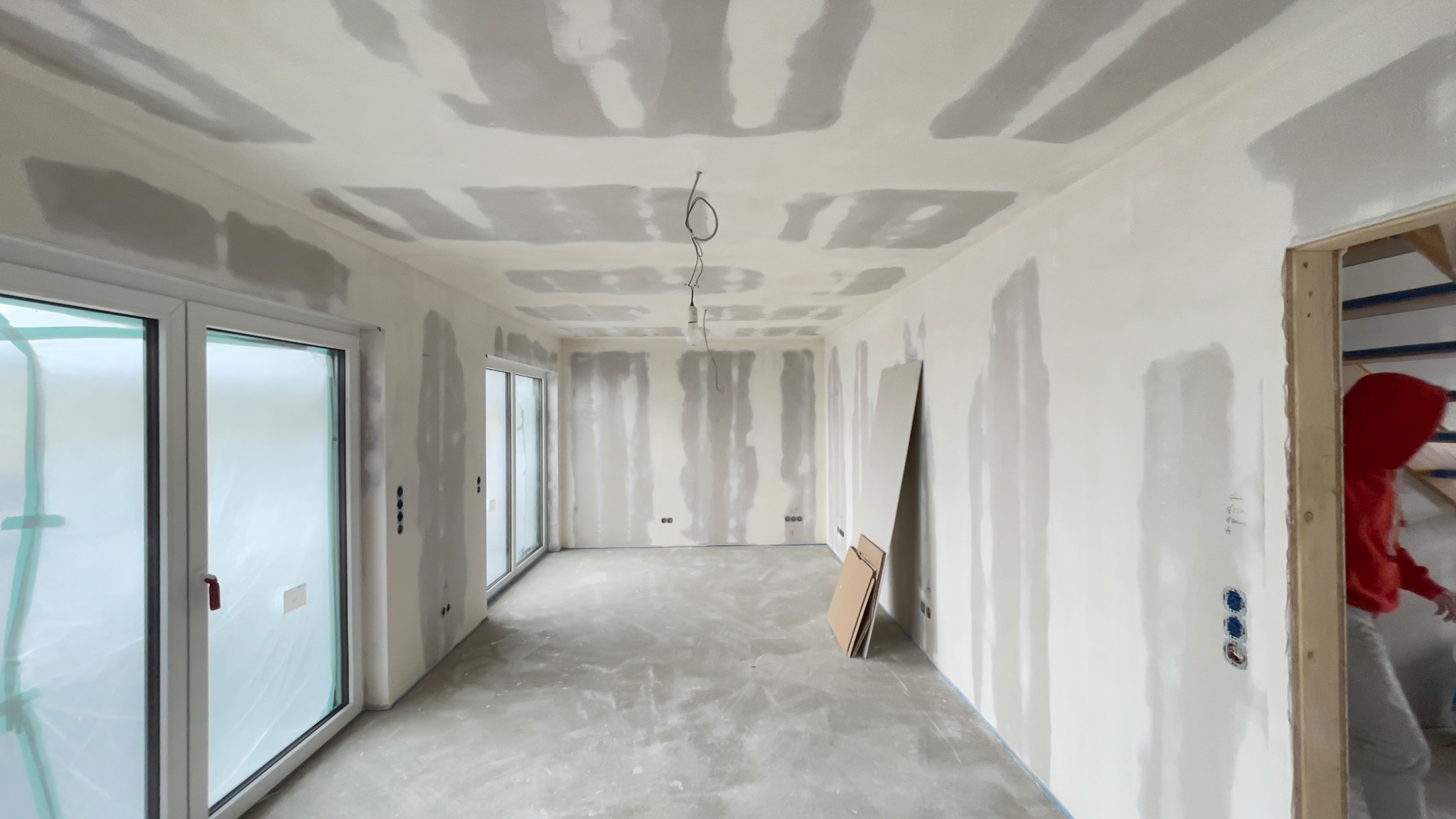 Wnętrze pokoju w trakcie prac przygotowawczych do malowania, widoczne ślady po szpachlowaniu na ścianach i suficie, dwa duże okna przesuwne z folią ochronną, wystające kable z sufitu, oraz osoba...