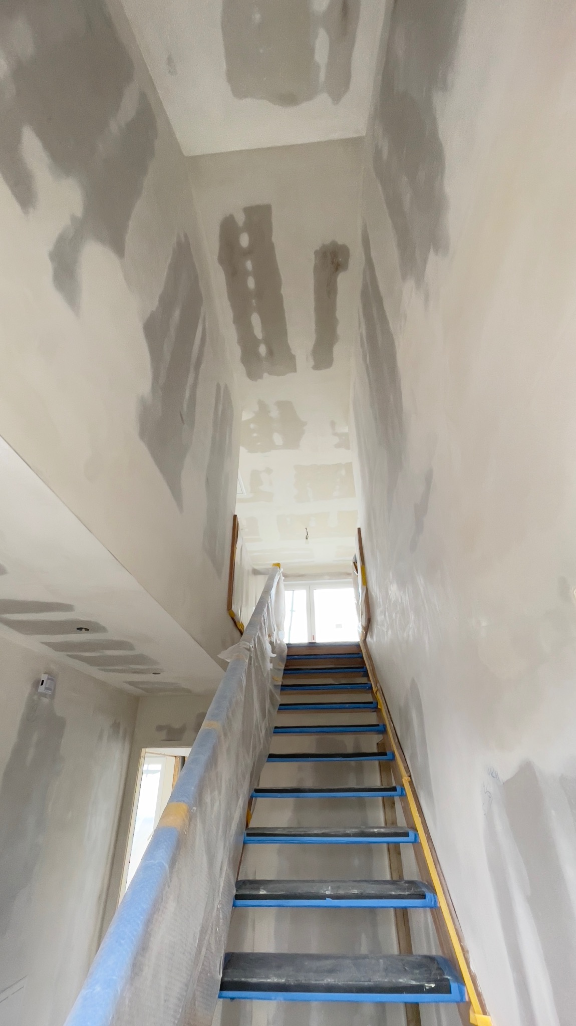 Klatka schodowa w trakcie przygotowania do malowania, zabezpieczona folią i taśmą malarską, ściany i sufit częściowo pokryte gładzią, widoczne nierówności i ślady po szpachlowaniu.