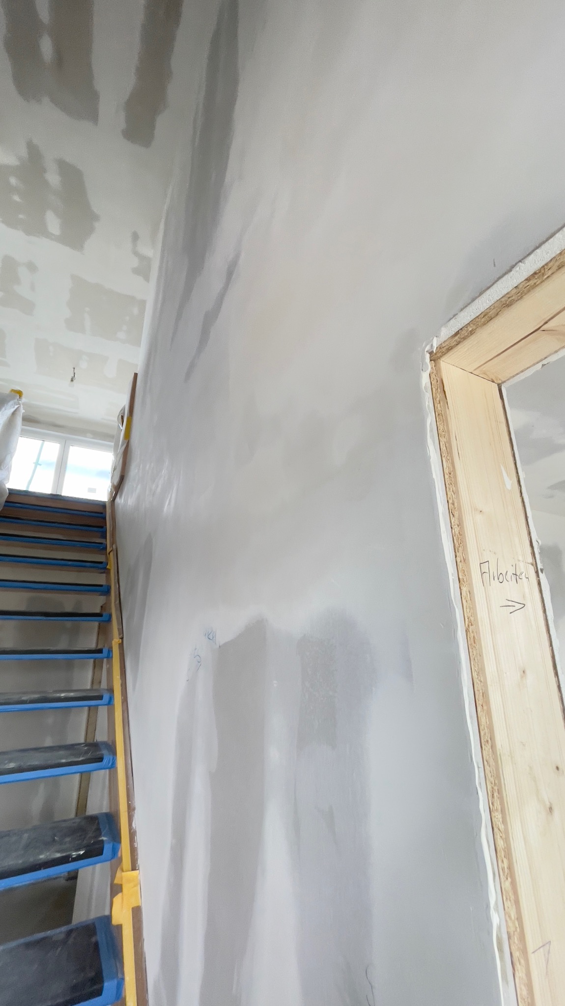 Ściana po nałożeniu pierwszej warstwy masy szpachlowej, widoczne ślady po szpachli, schody z niebieską taśmą zabezpieczającą oraz ościeżnica z napisem 'Flibeltest' i strzałką.