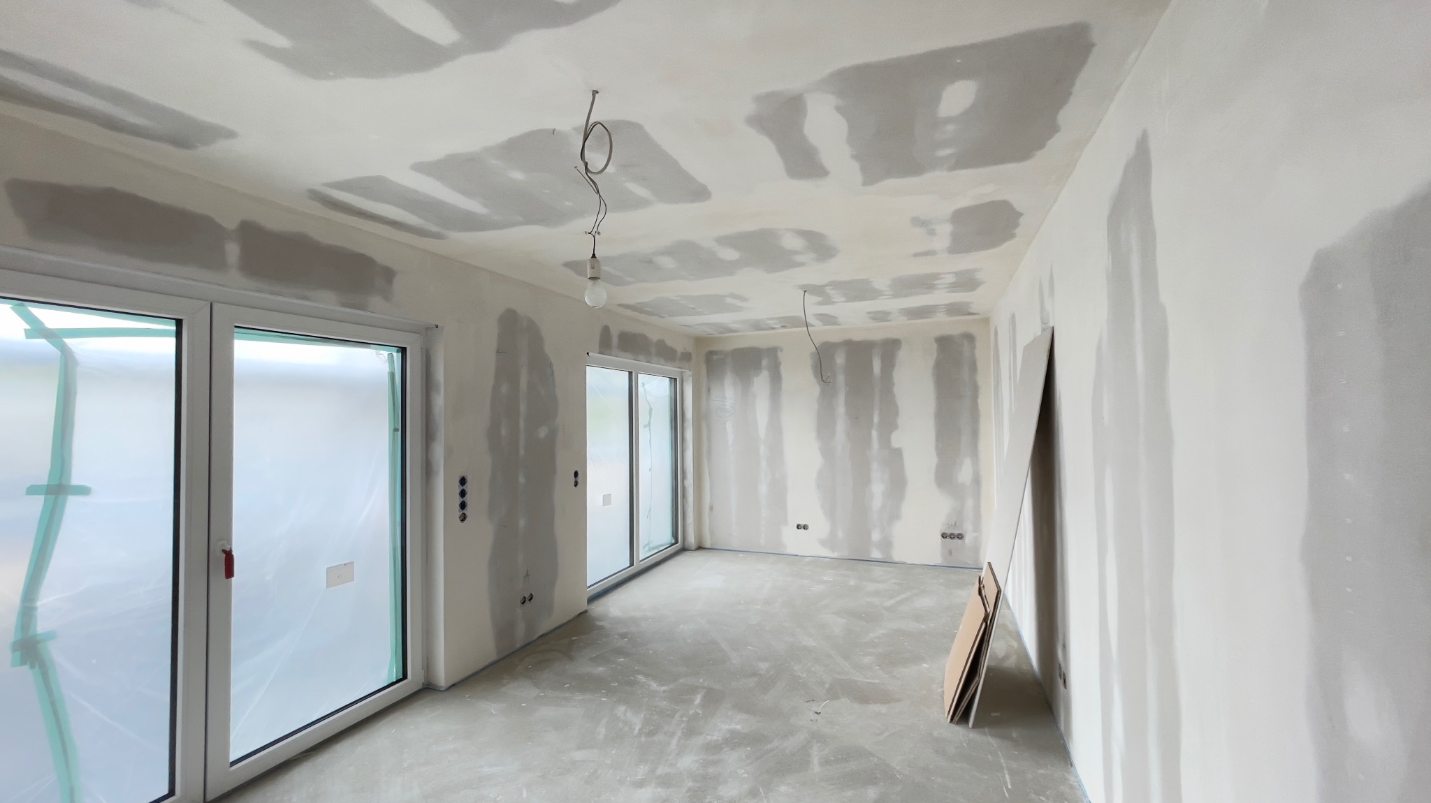 Pokój w trakcie prac przygotowawczych do malowania: ściany i sufit po szpachlowaniu, widoczne ślady łączeń płyt i nierówności, okna zabezpieczone folią, przewody oświetleniowe zwisają z sufitu.