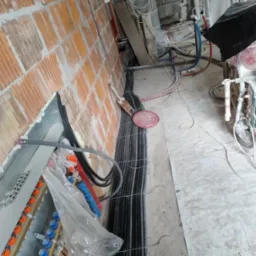 Wiązka przewodów elektrycznych w peszlach, prowadzona wzdłuż ściany z cegły dziurawki, z widoczną skrzynką rozdzielczą z niebieskimi i pomarańczowymi elementami, na podłodze pył budowlany.