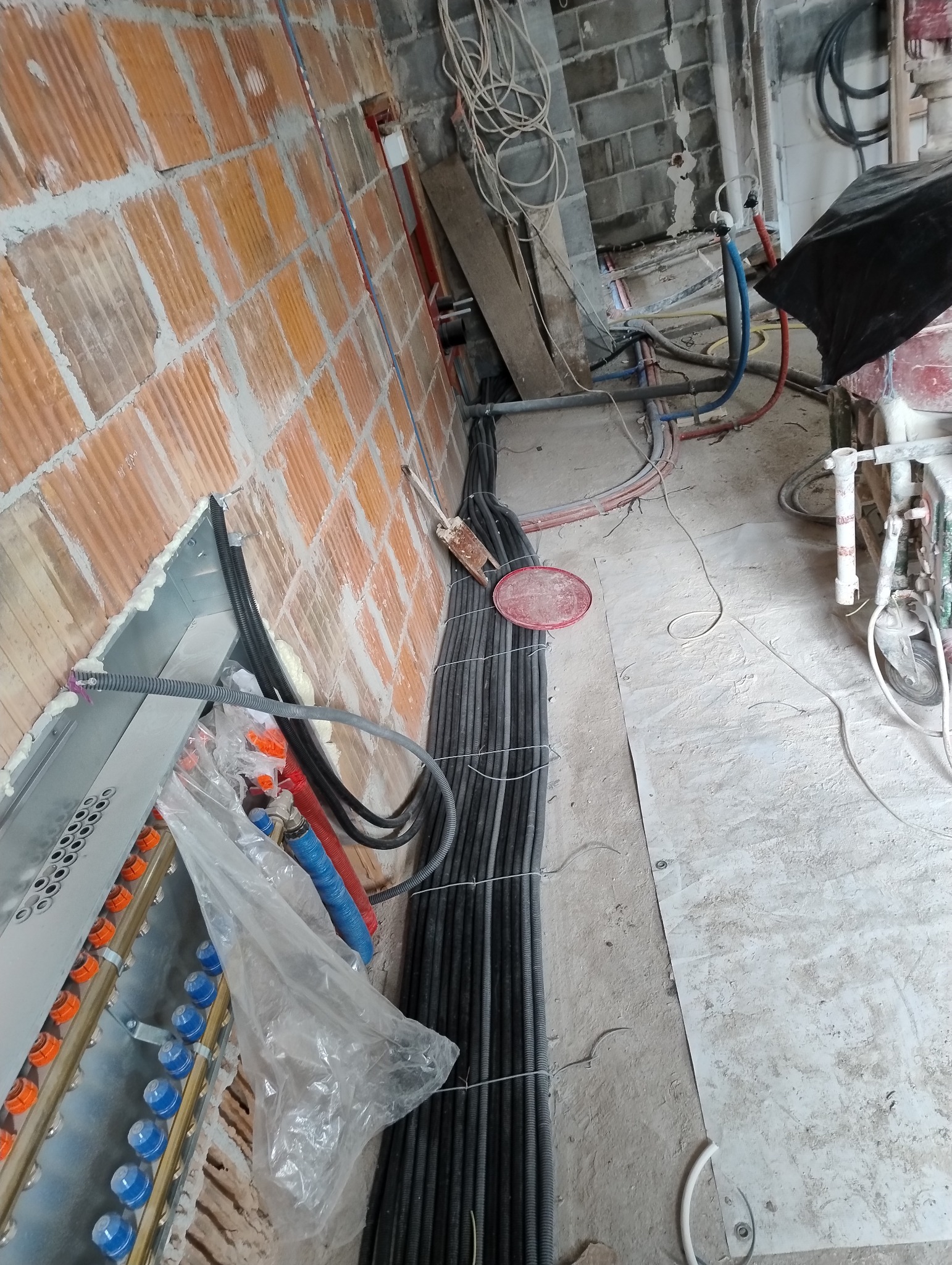 Wiązka przewodów elektrycznych w peszlach, prowadzona wzdłuż ściany z cegły dziurawki, z widoczną skrzynką rozdzielczą z niebieskimi i pomarańczowymi elementami, na podłodze pył budowlany.
