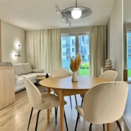 Jasne, przestronne wnętrze mieszkania z okrągłym stołem, krzesłami z beżową tapicerką, drewnianą podłogą i widokiem na budynek z balkonami za oknem.