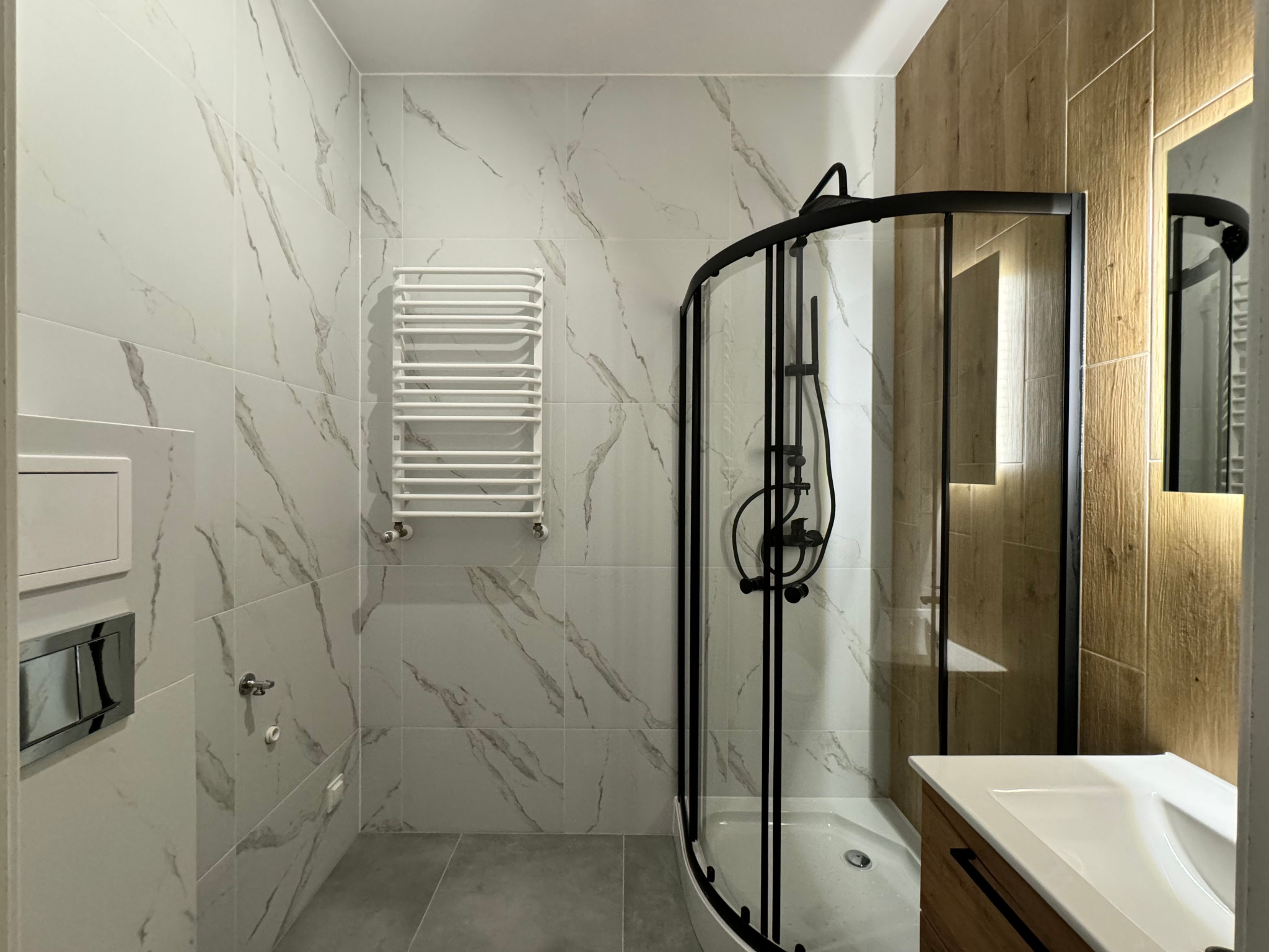 Nowoczesna łazienka z kabiną prysznicową z czarnymi profilami, grzejnikiem drabinkowym i płytkami imitującymi marmur na ścianach.