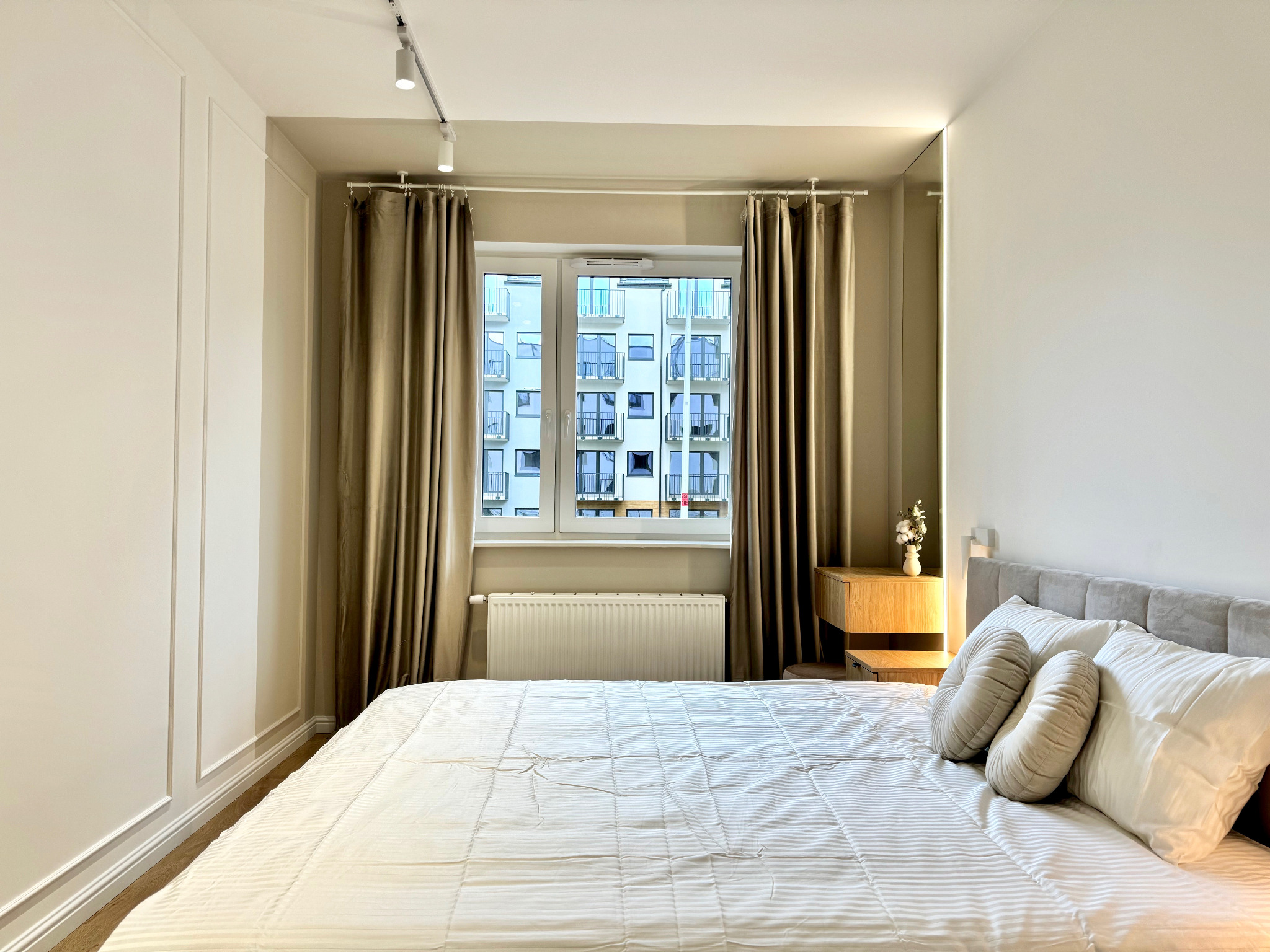 Jasna sypialnia z dużym oknem i beżowymi zasłonami, łóżko z szarą ramą i dekoracyjnymi okrągłymi poduszkami, ściana z dekoracyjnymi panelami.