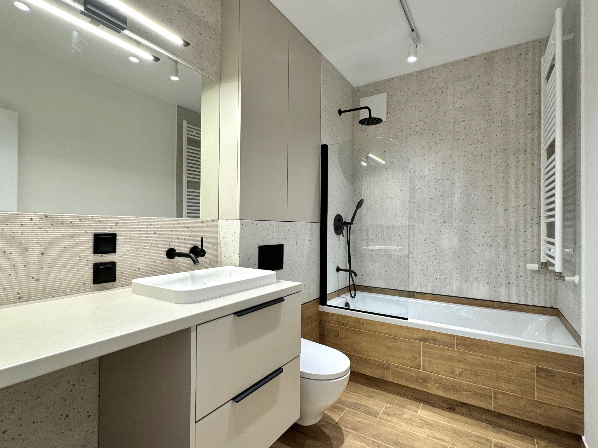 Nowoczesna łazienka z beżowymi szafkami pod umywalką, białą umywalką nablatową, czarną armaturą, wanną obudowaną drewnopodobnymi płytkami i mozaikową ścianą.