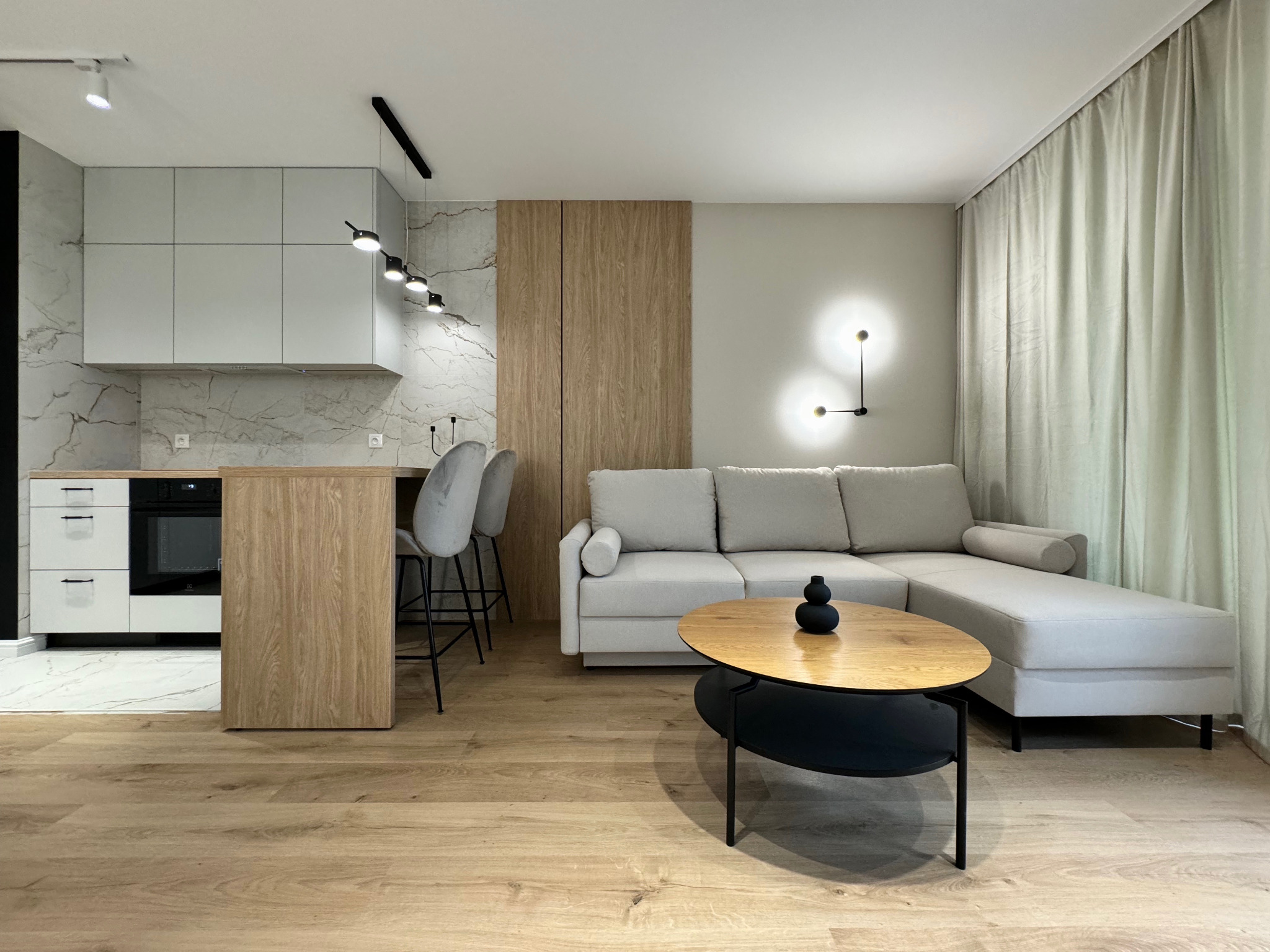 Nowoczesne wnętrze: salon z aneksem kuchennym, drewniana podłoga, szara sofa narożna, owalny stolik kawowy z drewnianym blatem, białe szafki kuchenne z marmurowym backsplash, oświetlenie punktowe...