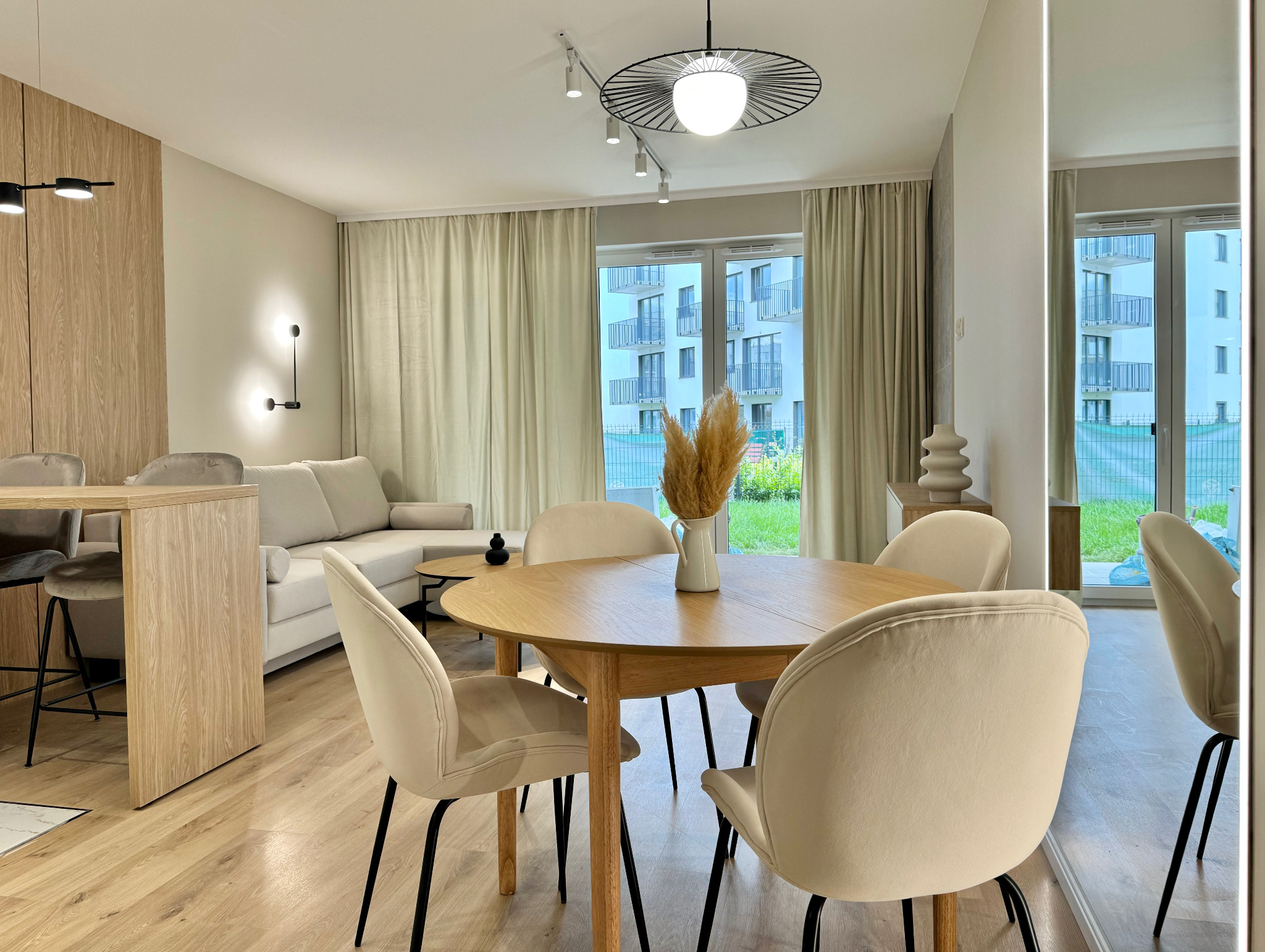 Jasne, przestronne wnętrze mieszkania z okrągłym stołem, krzesłami z beżową tapicerką, drewnianą podłogą i widokiem na budynek z balkonami za oknem.
