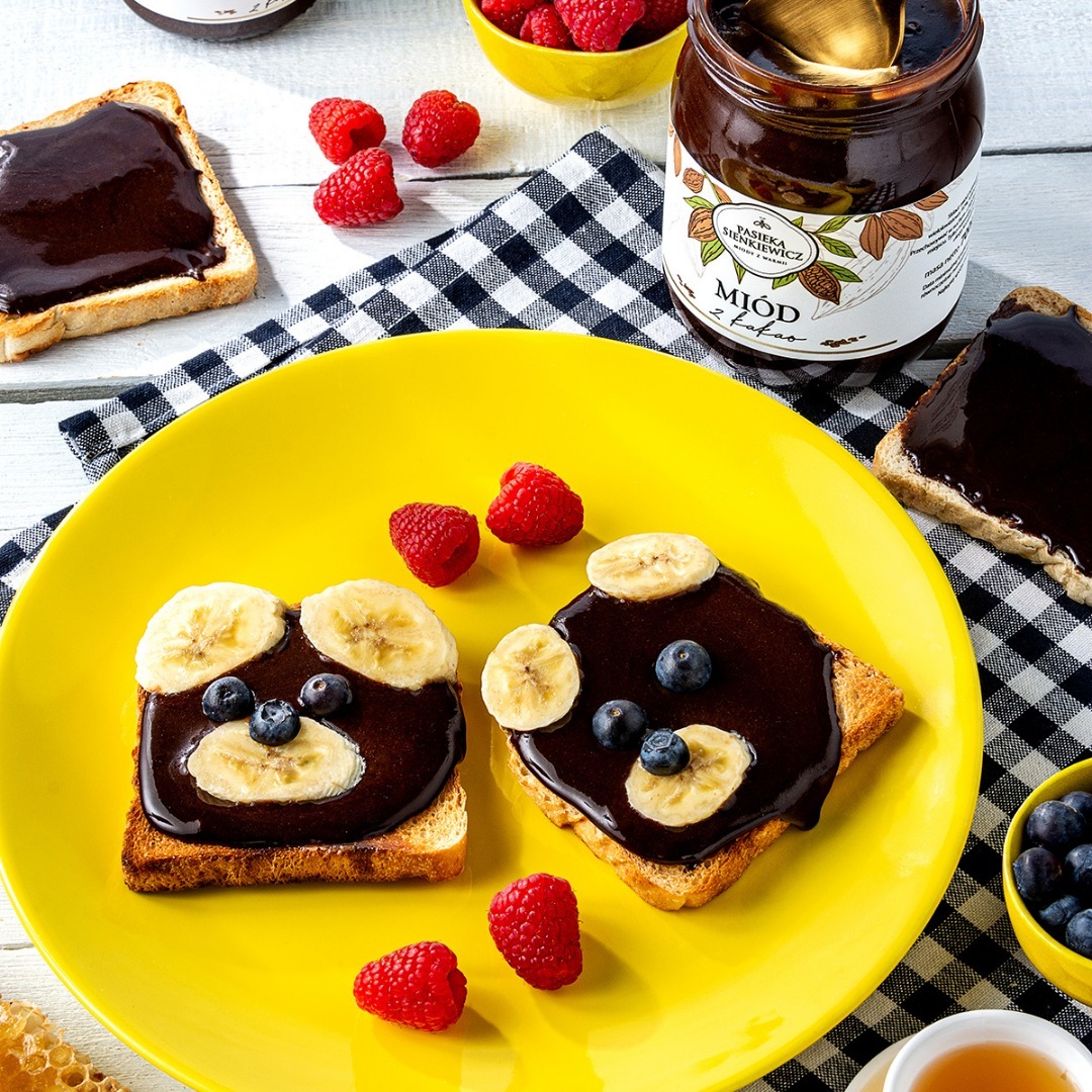 Tosty z kremem czekoladowym, bananami i borówkami na żółtym talerzu, ułożone w kształt misia, obok maliny, słoik kremu czekoladowego i tosty z samym kremem na tle czarno-białej ściereczki.
