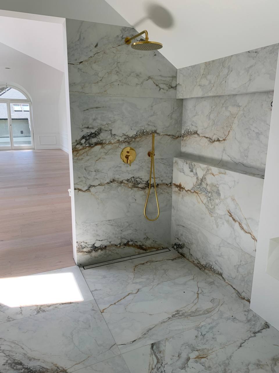 Nowoczesna kabina prysznicowa z marmurowymi płytkami i złotą armaturą, widok z zewnątrz na jasne, przestronne pomieszczenie z drewnianą podłogą.