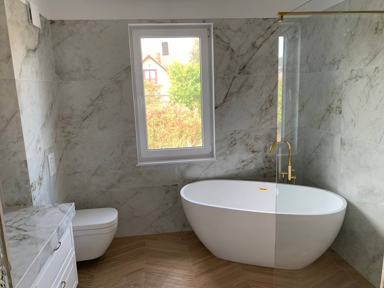 Nowoczesna łazienka z wanną wolnostojącą, złotą armaturą i marmurowymi płytkami ściennymi, widok na ogród przez okno.