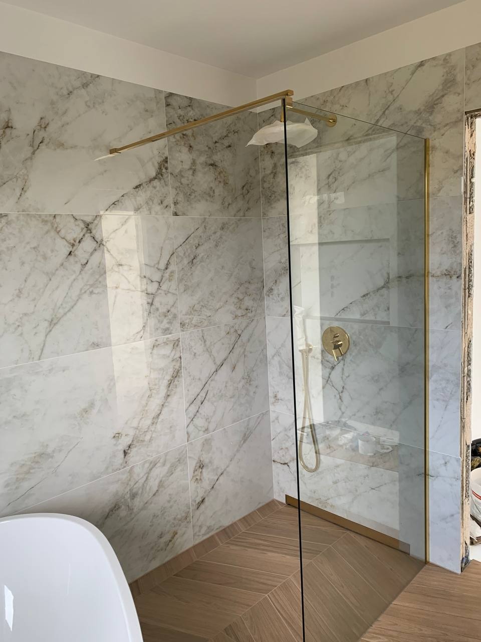 Wykończona łazienka z kabiną prysznicową ze złotymi detalami, marmurowe płytki na ścianach i drewnopodobna podłoga ułożona w jodełkę.