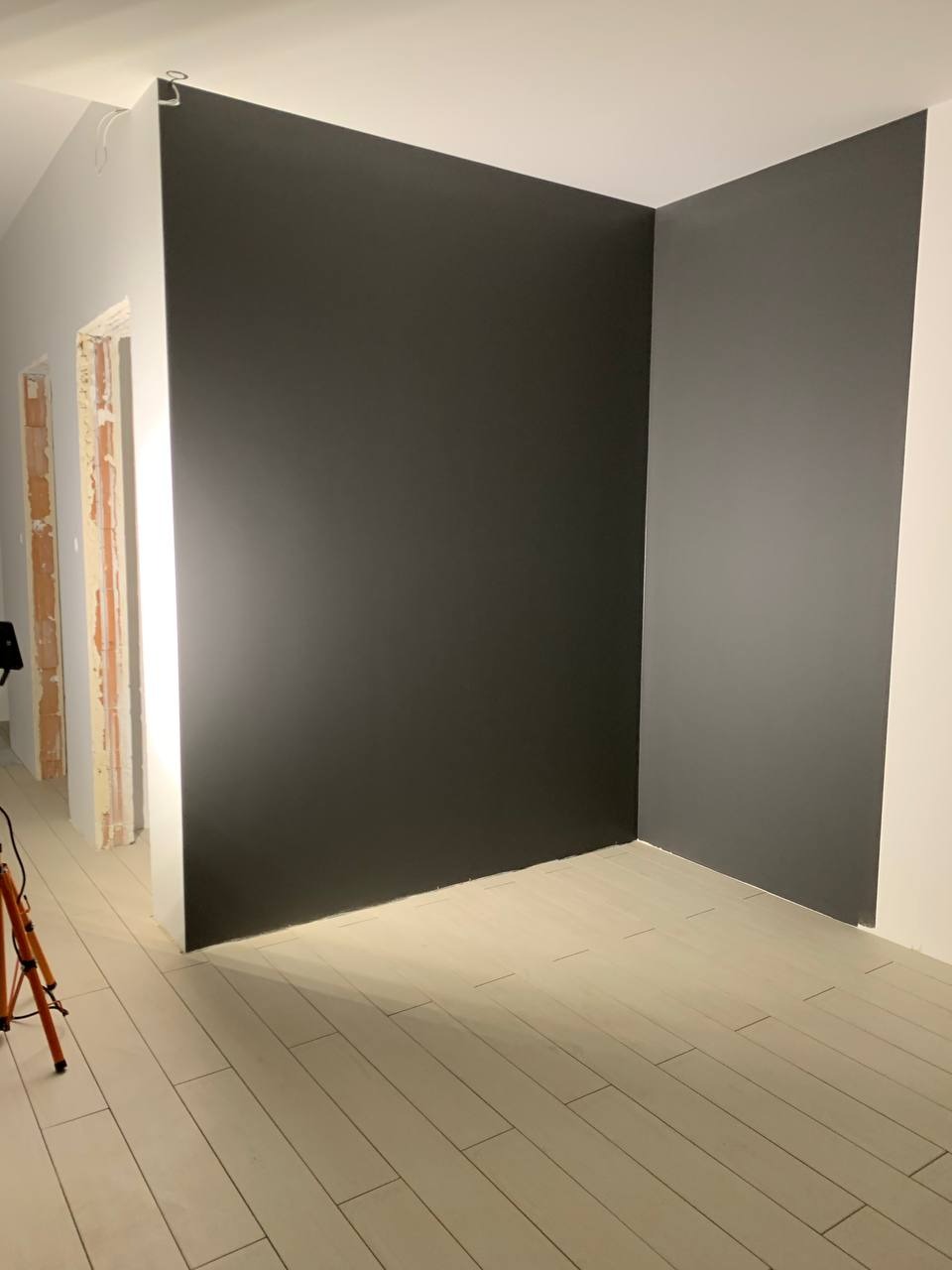 Świeżo pomalowane na ciemnoszaro ściany w rogu pokoju z jasną podłogą z paneli, widoczny fragment statywu oświetleniowego.