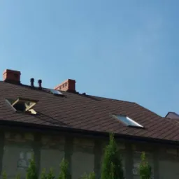 Brązowy dach z dachówki bitumicznej na budynku z ceglanym kominem, otwartym oknem dachowym i rynnami, na tle błękitnego nieba.