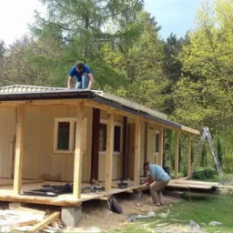 Dwóch mężczyzn pracuje nad budową drewnianego domku letniskowego z widoczną konstrukcją dachu i werandą, narzędzia rozłożone na podłodze, drabina oparta o ścianę, otoczenie leśne.