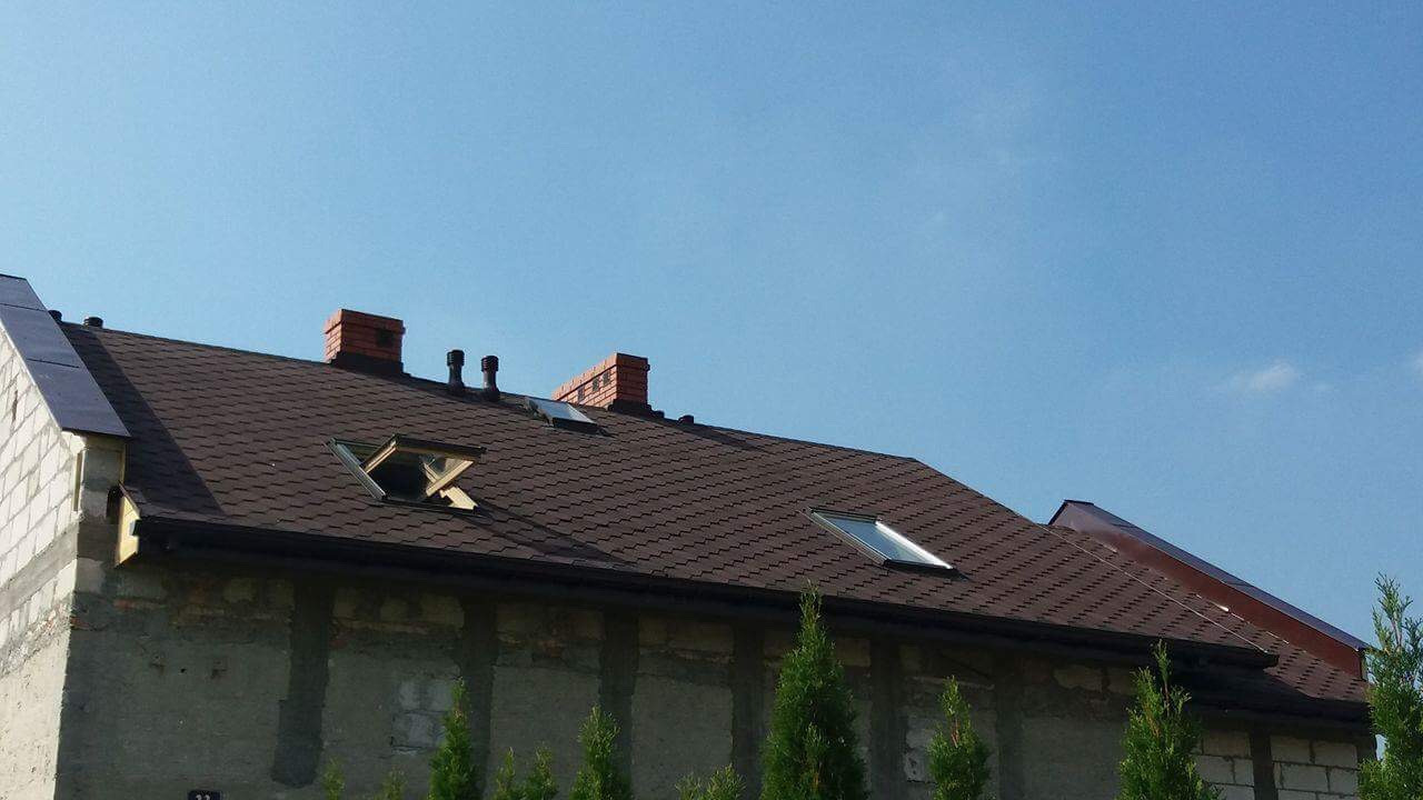 Brązowy dach z dachówki bitumicznej na budynku z ceglanym kominem, otwartym oknem dachowym i rynnami, na tle błękitnego nieba.