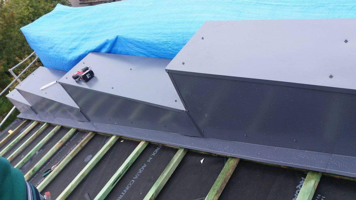 Szare, metalowe obróbki kominowe na dachu pokrytym membraną i łatami, zabezpieczone niebieską plandeką, narzędzia na górze.