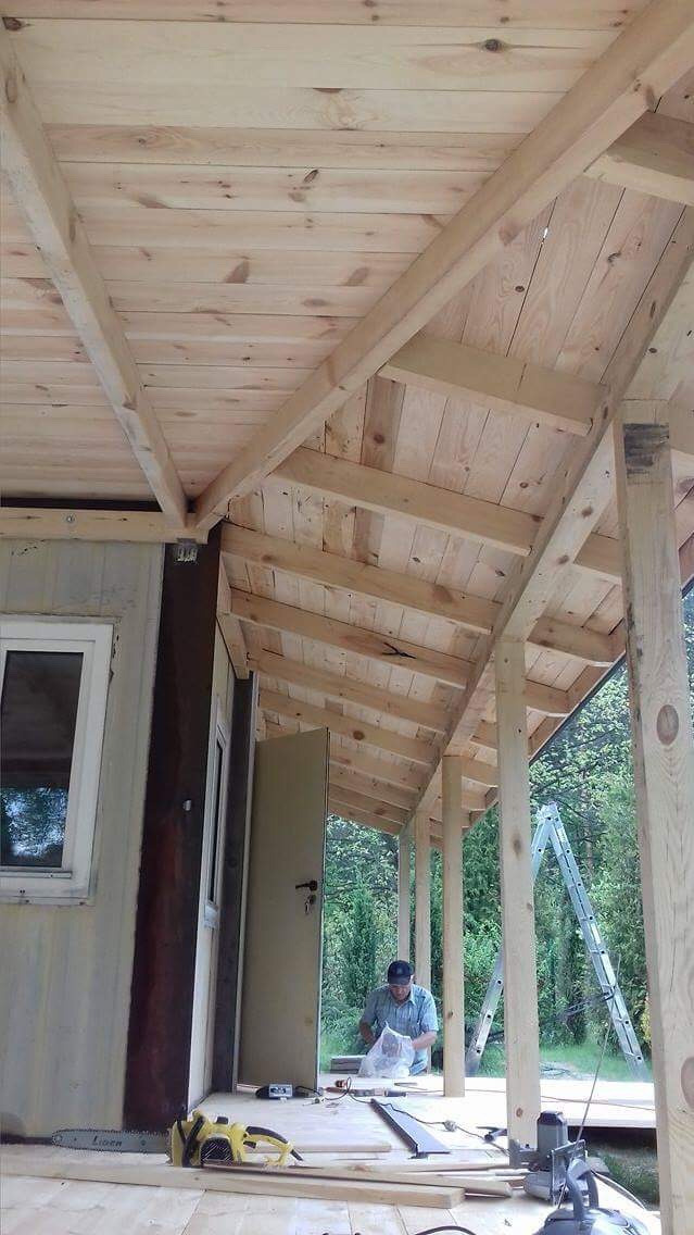 Konstrukcja drewnianej wiaty z widoczną więźbą dachową, deskowaniem i słupami nośnymi, w tle pracujący mężczyzna i drabina.