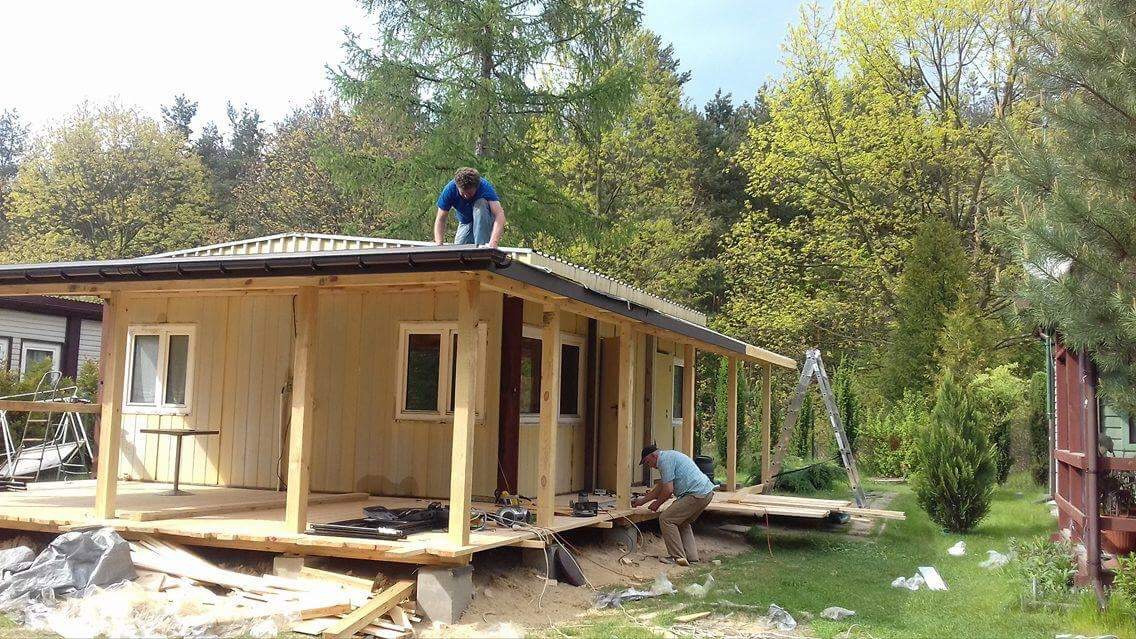 Dwóch mężczyzn pracuje nad budową drewnianego domku letniskowego z widoczną konstrukcją dachu i werandą, narzędzia rozłożone na podłodze, drabina oparta o ścianę, otoczenie leśne.