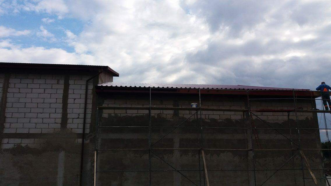 Budynek w trakcie prac dekarskich: robotnik na rusztowaniu montuje elementy orynnowania, widoczna surowa ściana z bloczków i fragment nowej, metalowej konstrukcji dachu.