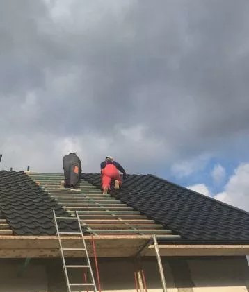 Dwóch dekarzy w kombinezonach roboczych montuje czarne blachodachówki na drewnianej konstrukcji dachu, widoczna drabina i pochmurne niebo.