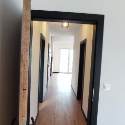 RN-BUD REMIGIUSZ ZDANOWICZ - Wnętrze korytarza z drewnianą podłogą, widok przez otwarte drzwi z czarną ramą, w tle widoczne kolejne drzwi i okno wpadające światło dzienne.