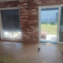 RN-BUD REMIGIUSZ ZDANOWICZ - Układanie parkietu w jodełkę na podkładzie z folii, widoczny młotek i fragment ściany z cegły dekoracyjnej, okno z roletą i przesuwne drzwi balkonowe.