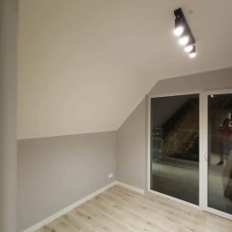 RN-BUD REMIGIUSZ ZDANOWICZ - Pokój na poddaszu po remoncie: ściany pomalowane na dwa odcienie szarości, jasna podłoga z paneli, czarne oświetlenie sufitowe i duże okno balkonowe z widokiem na zewnątrz.