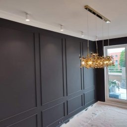 RN-BUD REMIGIUSZ ZDANOWICZ - Eleganckie wnętrze po malowaniu, ciemnoszara ściana z dekoracyjnymi panelami, nowoczesny złoty żyrandol, podłoga zabezpieczona folią malarską, widok na balkon z pracującym mężczyzną.