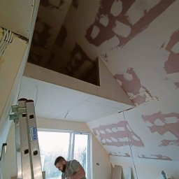 RN-BUD REMIGIUSZ ZDANOWICZ - Prace wykończeniowe poddasza: montaż płyt gipsowo-kartonowych na skosach, widoczny pracownik z płytą, drabina Krause i fragment okna.