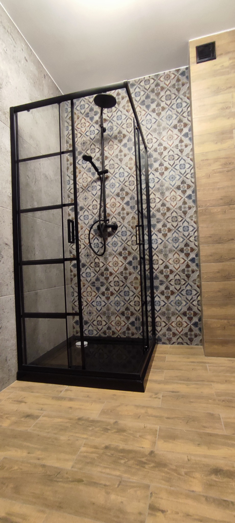 Kabina prysznicowa z czarnymi profilami i deszczownicą w wykończonej łazience, widoczne płytki imitujące drewno na podłodze i wzorzyste płytki na ścianie.