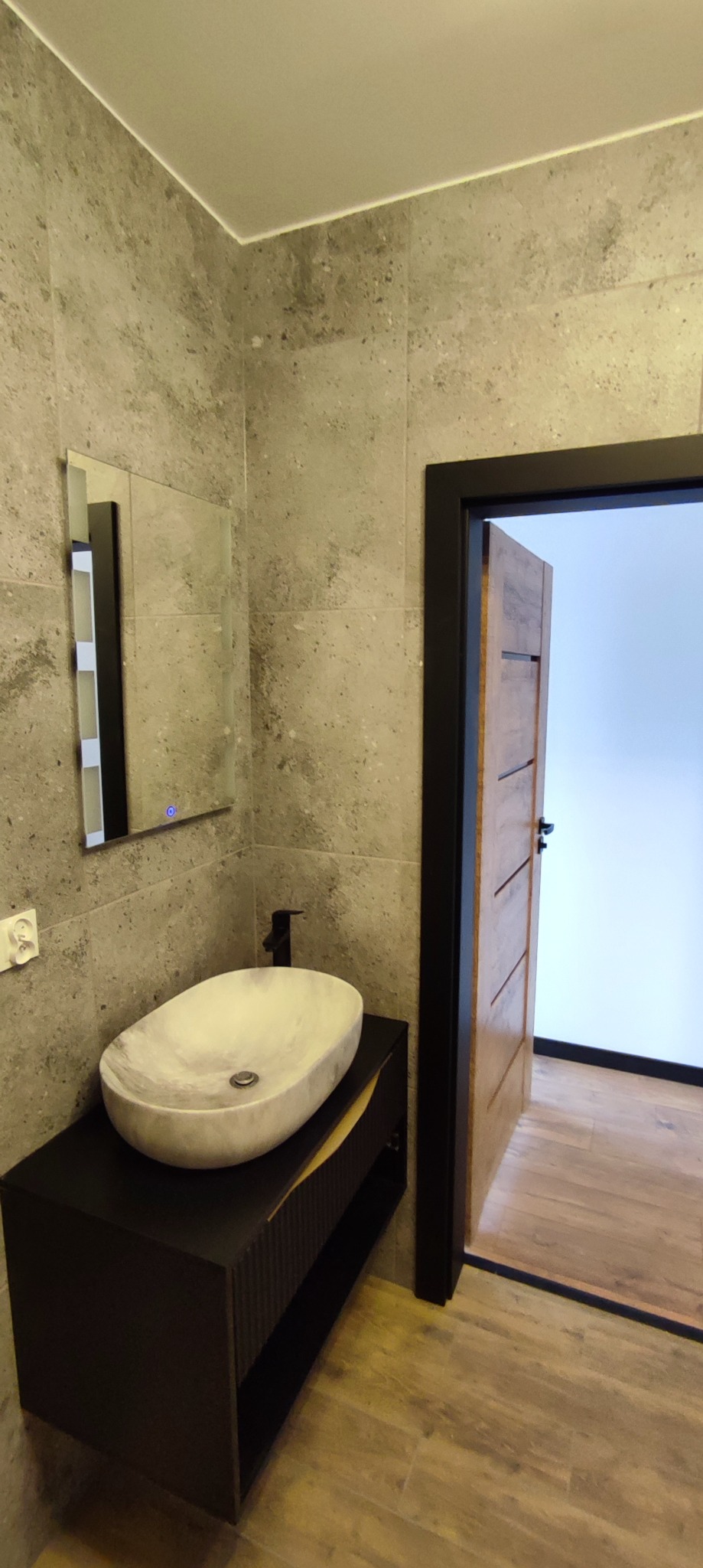 Nowoczesna łazienka z umywalką nablatową z marmuru, lustrem z podświetleniem LED, szarą płytką imitującą kamień na ścianach oraz drewnianymi panelami podłogowymi i drzwiami z czarną ościeżnicą.