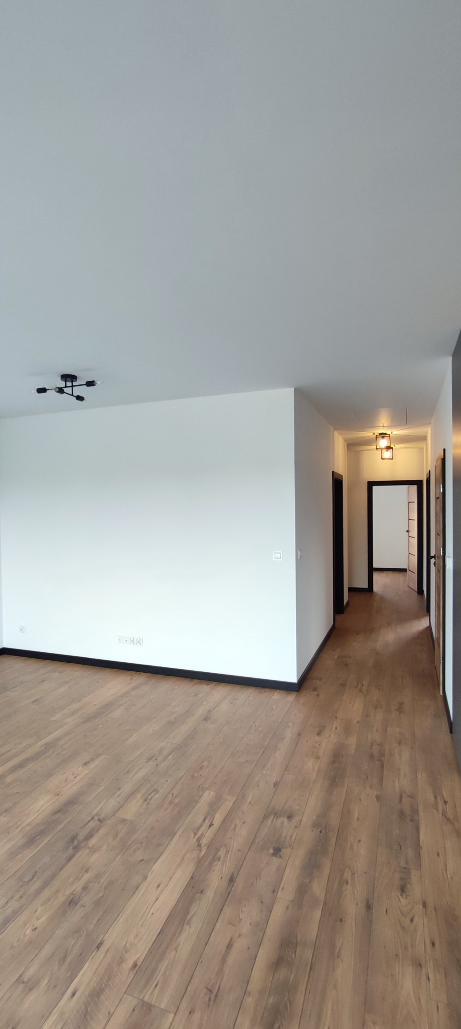 Jasne, przestronne wnętrze po remoncie w Śremie z drewnianą podłogą, białymi ścianami i czarnymi listwami przypodłogowymi, widok na korytarz z otwartymi drzwiami.