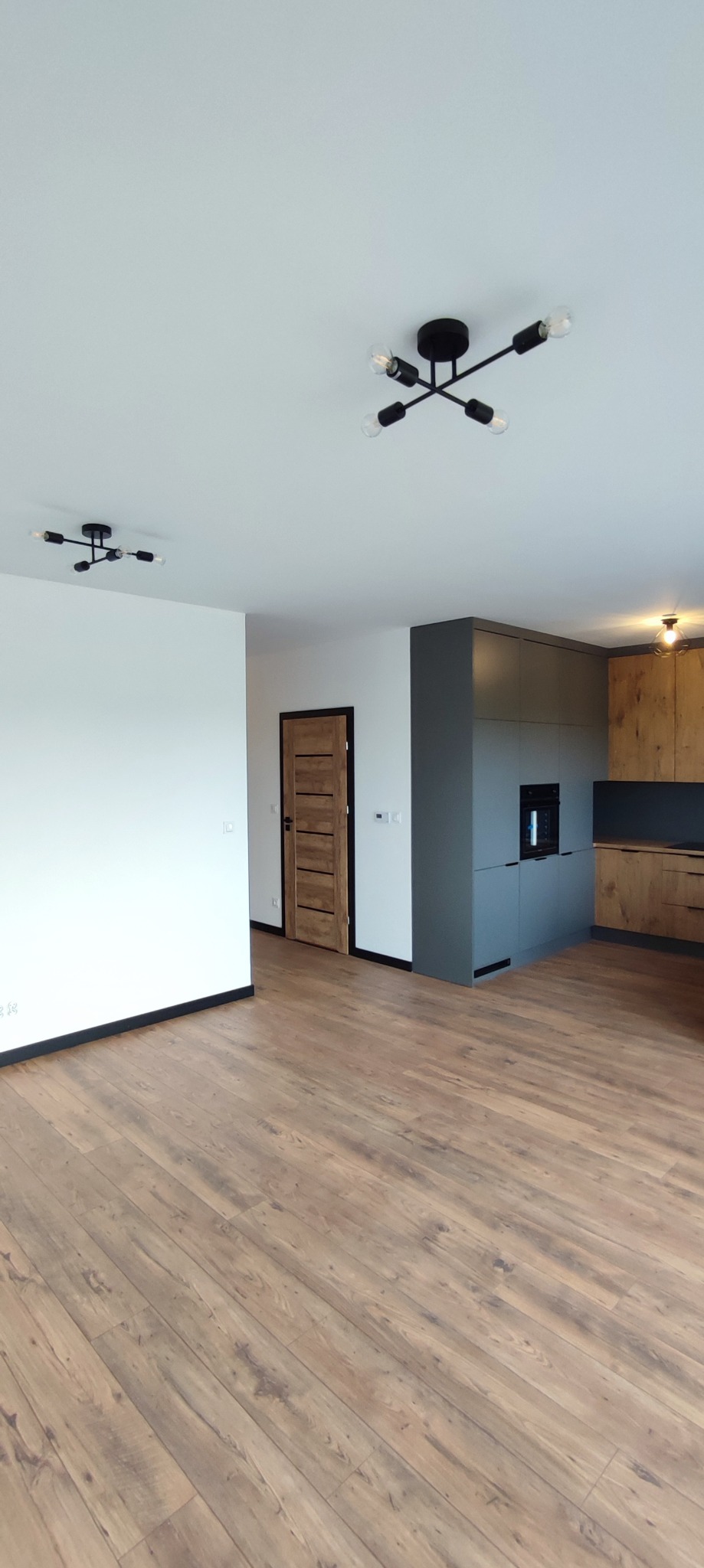 Nowoczesne wnętrze mieszkania z drewnianą podłogą, ciemnoszarą zabudową kuchenną i drewnianymi frontami szafek, widoczne dwie designerskie lampy sufitowe w stylu industrialnym.