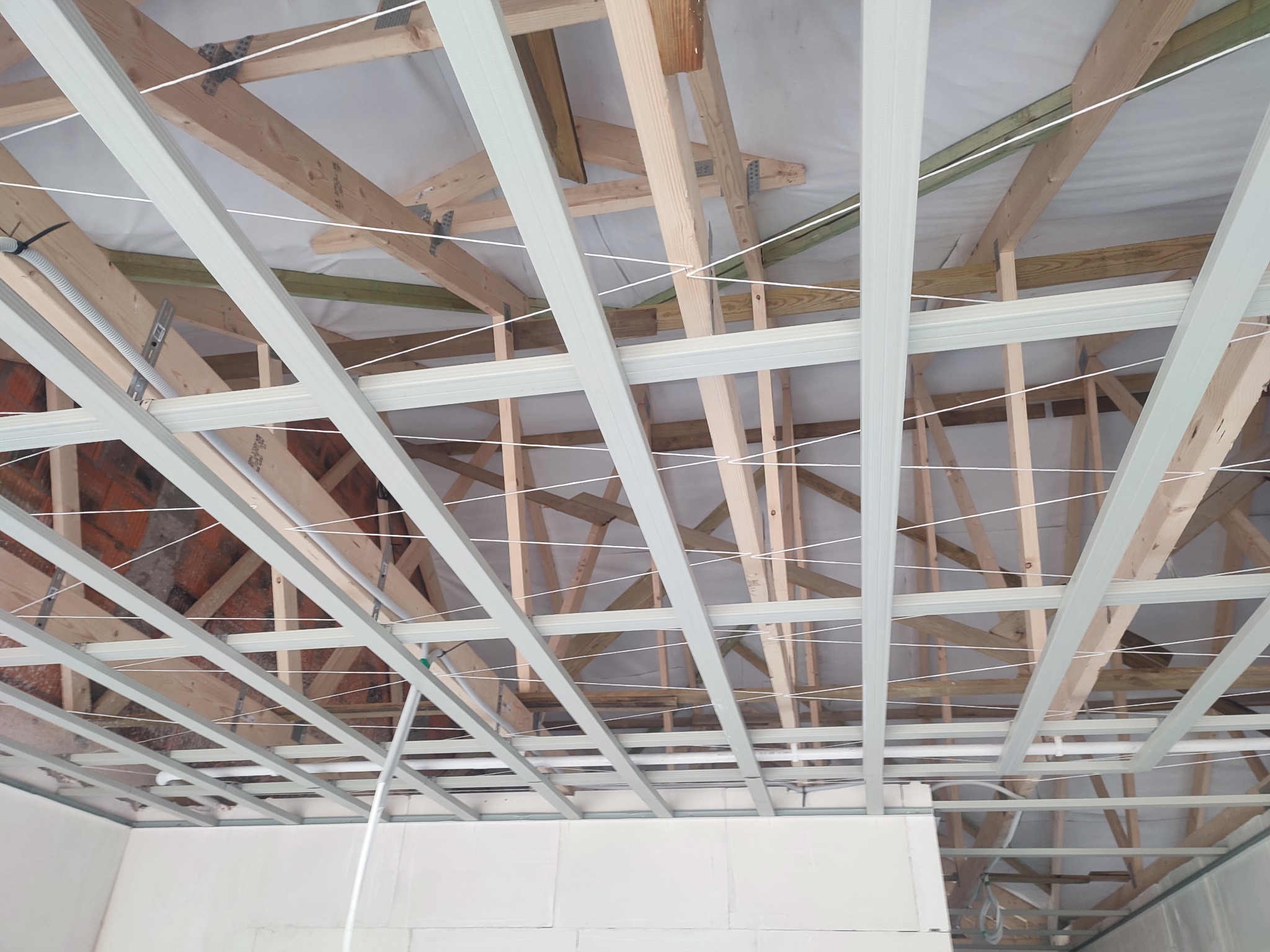 Konstrukcja sufitu podwieszanego z drewnianymi belkami stropowymi w tle, widoczne profile metalowe i białe sznurki do poziomowania.