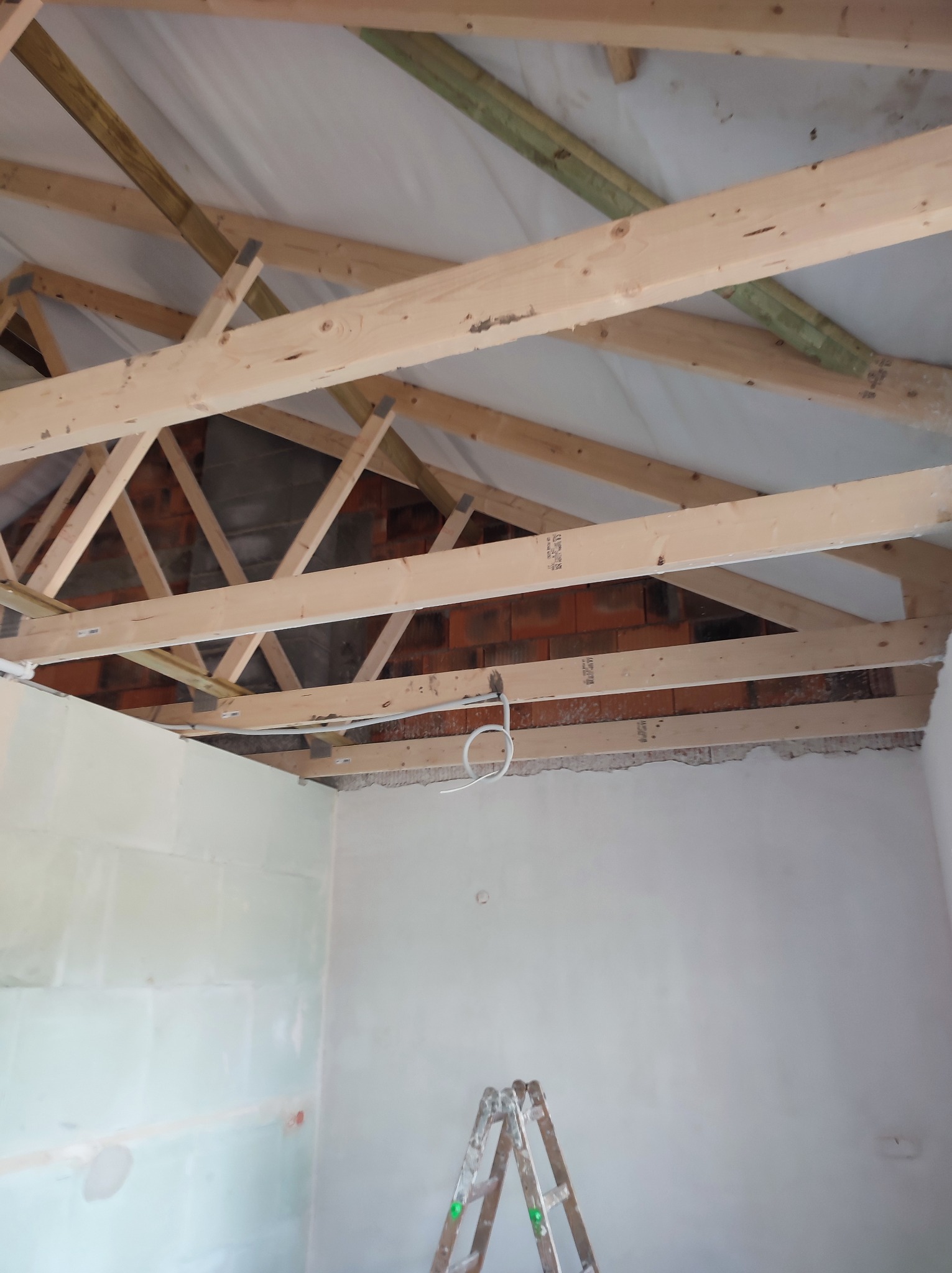 Surowy stan poddasza z widoczną konstrukcją drewnianą dachu, ścianami w trakcie przygotowania do wykończenia i drabinką malarską w rogu pomieszczenia.