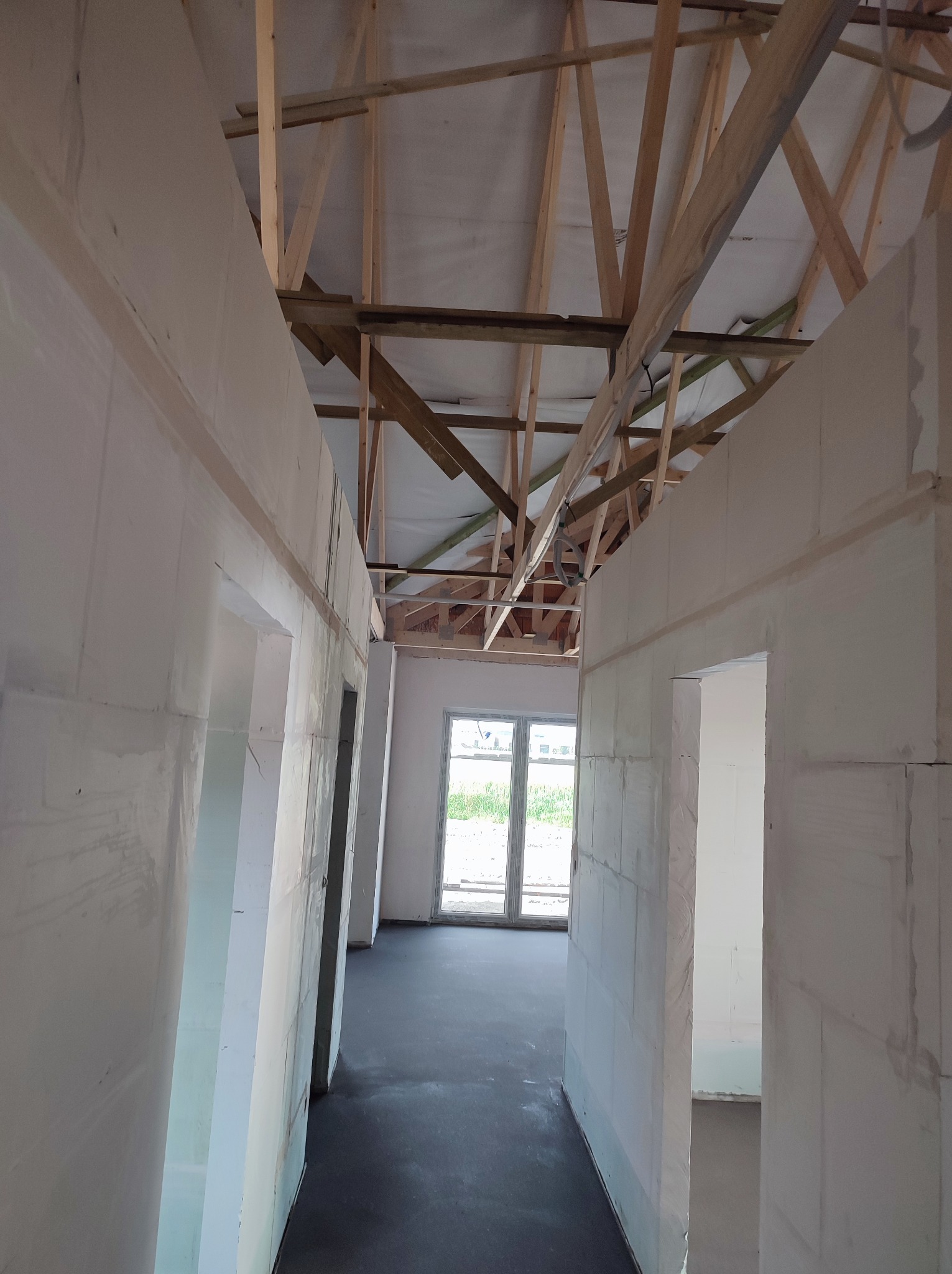 Wnętrze w trakcie budowy w Śremie, widoczne białe ściany z betonu komórkowego, surowa konstrukcja dachu z drewnianych belek oraz czarna podłoga wylewka.
