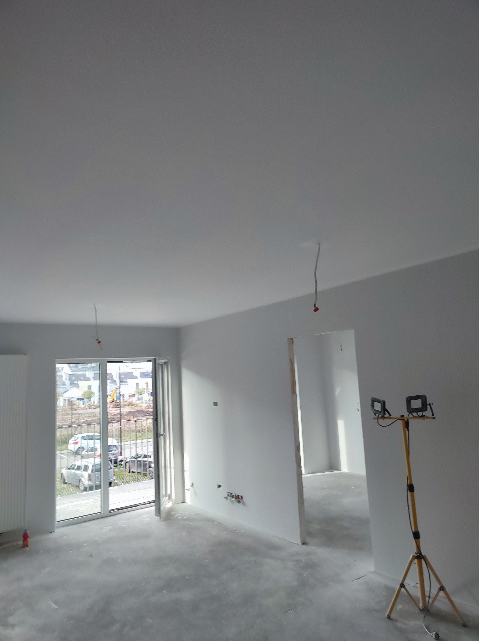Wnętrze w trakcie remontu: białe ściany, surowy beton na podłodze, otwarte drzwi balkonowe z widokiem na osiedle, lampa budowlana na statywie i wystające kable z sufitu.