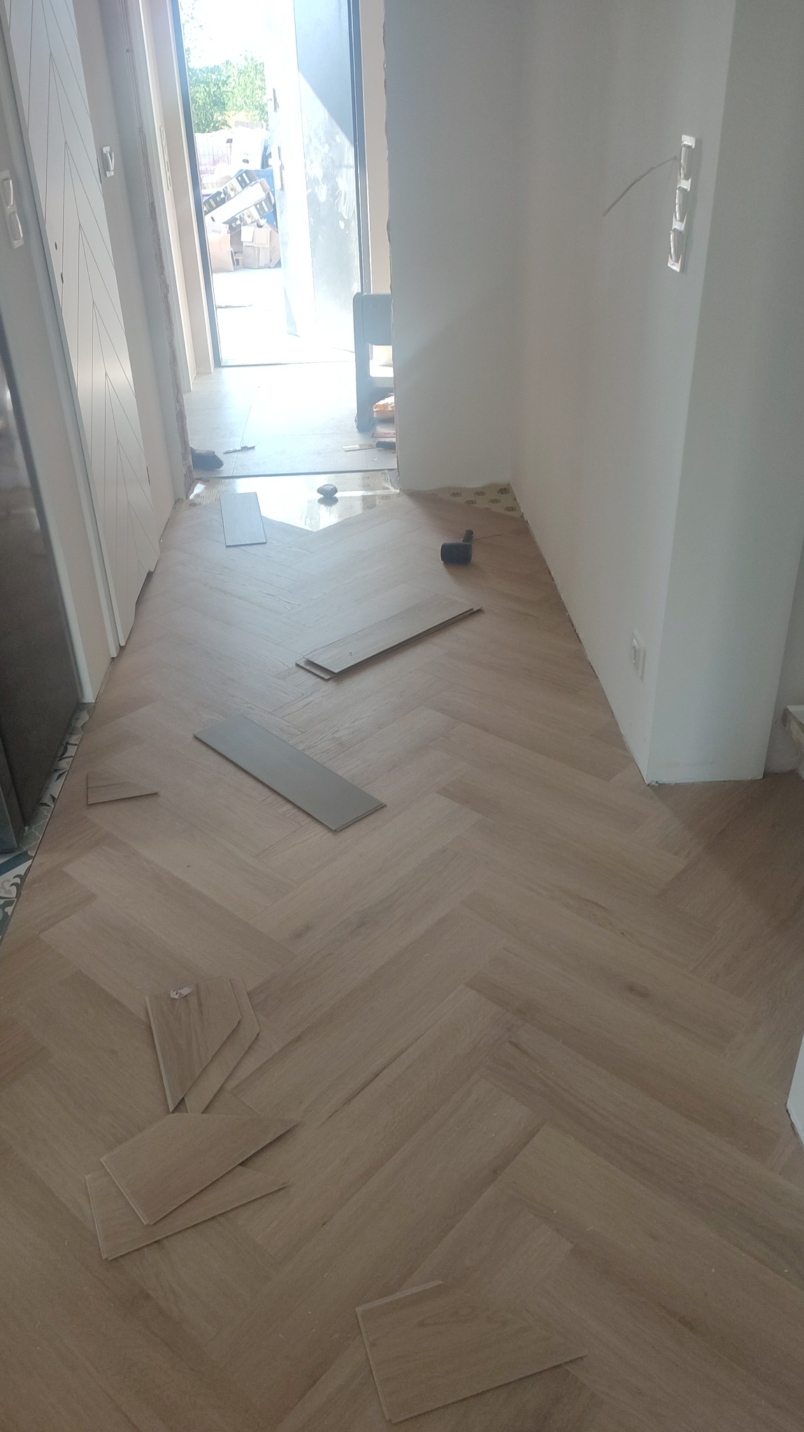 Fragment podłogi z ułożonymi panelami w jodełkę, kilka paneli leży luzem, widoczny młotek gumowy i listwa przypodłogowa