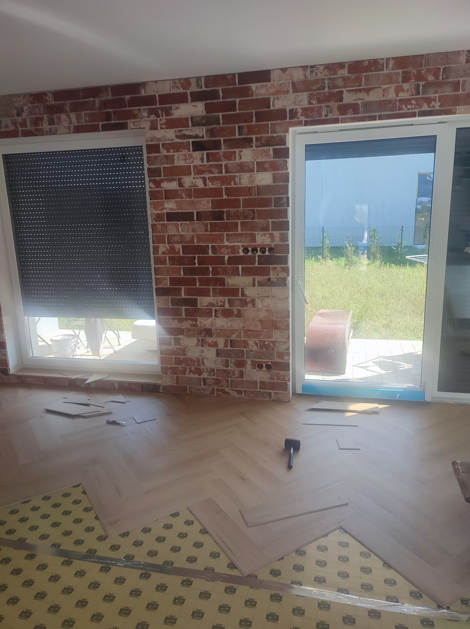 Układanie parkietu w jodełkę na podkładzie z folii, widoczny młotek i fragment ściany z cegły dekoracyjnej, okno z roletą i przesuwne drzwi balkonowe.